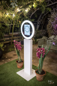 Open air white photo booth with mirror faceplate and custom welcome screen surrounded by plants